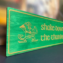 Load image into Gallery viewer, This Shake Down the Thunder wall art is laser cut and engraved and features a blue or green stained background with birch wood letters, accents and the Notre Dame Leprechaun. The art is 30" x 10" to make it perfect for your dorm room, apartment, or home!
