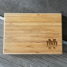 Load image into Gallery viewer, Wooden cheese board with a Notre Damelogo on a textured surface
